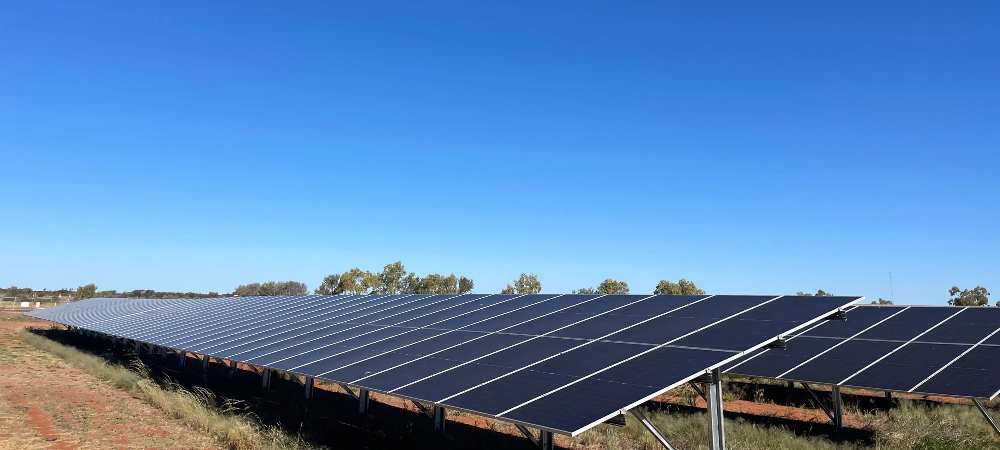 A long row of ground-mounted solar panels on red earth, with trees on the horizon under a cloudless blue sky.